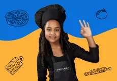 A young female kid chef somewhere between the ages of 7 and 9 with her arm up in the OK hand gesture wearing a black baker's cap, a black long-sleeved shirt and a black apron with the Vaughan Studios & Event Space logo on the front. 