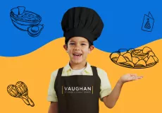 A young male kid chef somewhere between the ages of 7 and 9 smiling with an open-mouthed smile straight at the camera wearing a black baker's cap, yellow short-sleeved polo shirt with a white t-shirt underneath, and a black apron with the Vaughan Studios & Event Space logo on the front. 