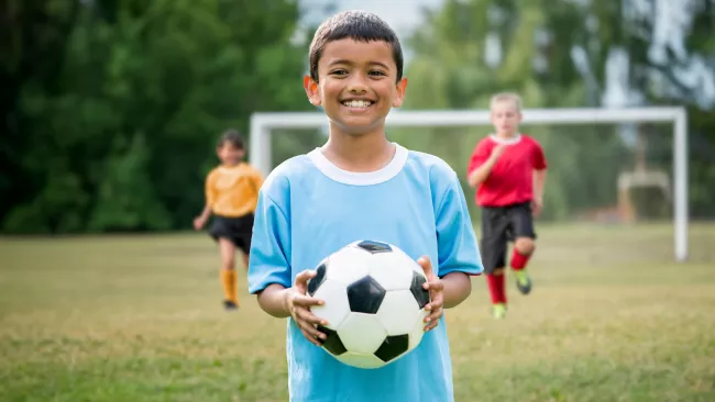 Little boy holding a soccer ball on the field with two kids running behind him