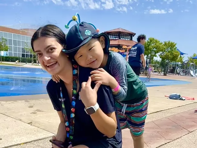 A smiling summer camp counsellor with a young boy hugging her sholders from behind.