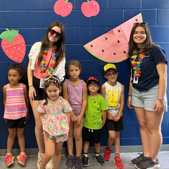 Two camp counsellors posing with five young campers in front of a blue wall with fruit cutouts.