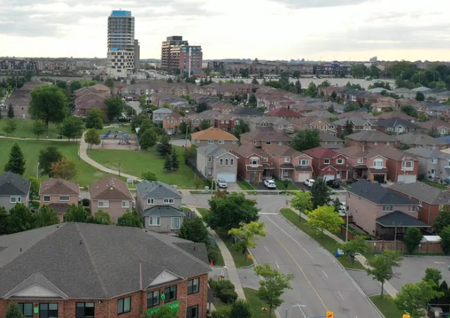 An elevated photo of a neighborhood including houses on, a path leading through greenspace and a few towers in the background.